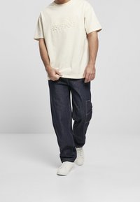 Krämfärgad t-shirt med präglad "SOUTHPole"-logga, ihop med mörkblå denimjeans med sidoficka och vita sneakers.