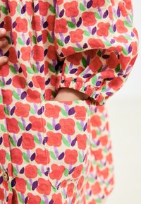 Skjorte i beige med blomsterprint, der viser røde blomster, grønne blade og lilla detaljer. Med en lomme og indsnørede ærmer.