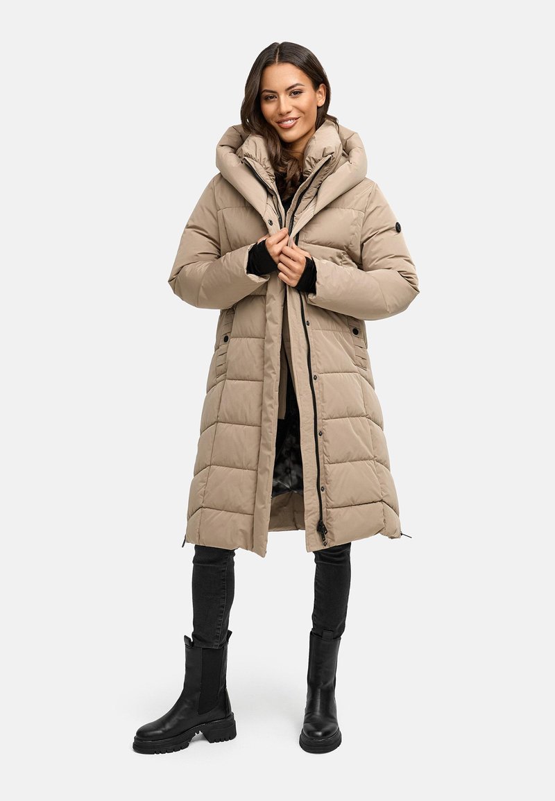 Giacca puffer beige con colletto alto e tasche abbottonate, caratterizzata da un design trapuntato e chiusura con zip, indossata sopra jeans neri e stivali.