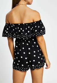 Kvinna klädd i en svart off-shoulder jumpsuit med stora och små vita prickar, sedd från ryggen mot en enfärgad bakgrund.
