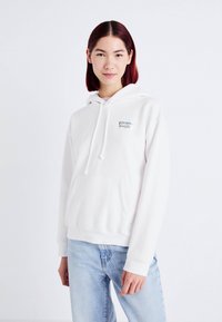 Vit huvtröja i mjukt material, med en framficka och en färgglad broderad logotyp på vänster sida.