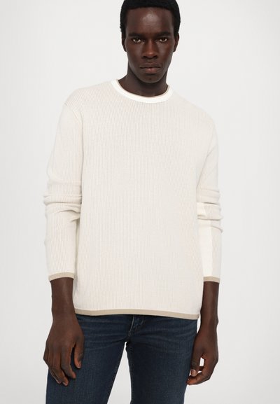 Weißer gerippter Strickpullover mit langen Ärmeln und Rundhalsausschnitt. Die Bündchen und der Saum sind mit einem hellbeigen Akzent versehen. Getragen mit dunkelblauen Jeans.