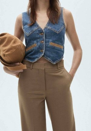 Žena v bezrukávové džínové vestě a hnědých kalhotách s vysokým pasem, držící v jedné ruce hnědý klobouk, s druhou rukou v kapse.