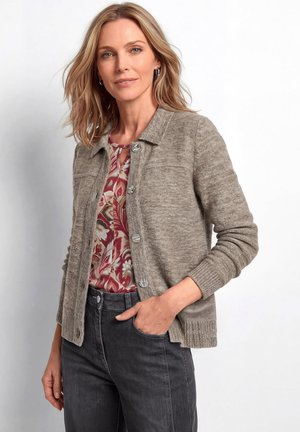 Frau mit schulterlangen blonden Haaren, die eine beige, geknöpfte Strickjacke über einem geblümten Top und dunklen Jeans trägt und mit einer Hand in der Tasche steht.