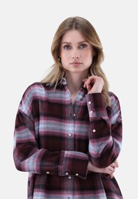 Junge Frau mit blonden Haaren, die ein karierter Button-Up-Hemd in Bordeauxrot und Rosa trägt, posiert mit einer Hand am Kinn und der anderen verschränkt.