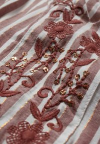 Tissu rayé aux nuances roses et blanches, présentant une broderie florale complexe en accents rouges et dorés, mettant en valeur la texture et les détails.