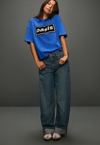 Koningsblauw T-shirt met zwart "oasis"-logo, gecombineerd met wijde blauwe jeans. Grijs-witte schoenen contrasteren met de outfit.