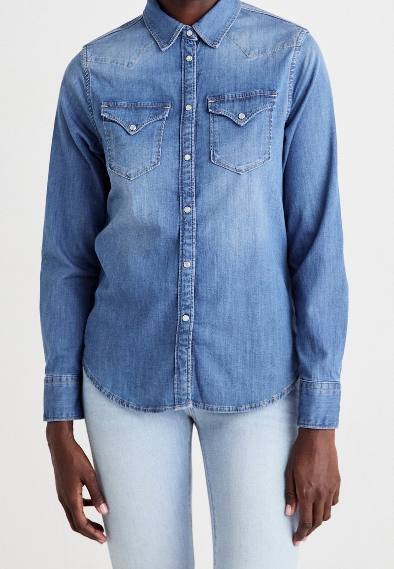 Homme portant une chemise en denim bleu clair avec des boutons-pression et deux poches poitrine associée à un jean clair sur fond uni.
