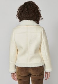 Krém színű báránybőr dzseki texturált felülettel, nagy gallérral, valamint szőrmével bélelt mandzsettákkal és alsó résszel. A hátulról készült nézet kiemeli a varrási részleteket.