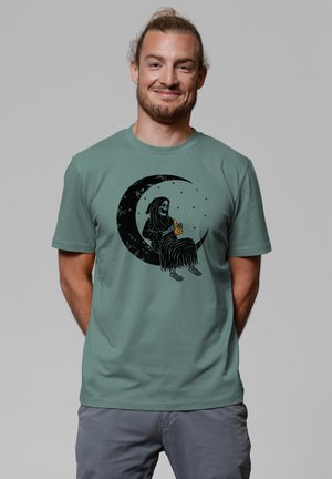 Green cotton t-shirt featuring a black graphic of a figure with long hair sitting on a crescent moon, surrounded by stars.