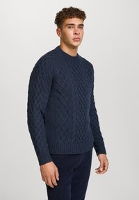 Granatowy sweter z dzianiny o fakturowanym wzorze w warkocze, okrągłym dekolcie oraz ściągaczach przy rękawach i na dole. Noszony przez modela na neutralnym tle.
