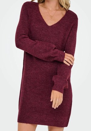 Femme portant une robe-pull tricotée bordeaux à manches longues et col en V, avec une main reposant sur le bras opposé, sur un fond uni.