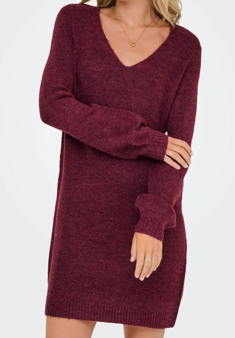 Femme portant une robe-pull tricotée bordeaux à manches longues et col en V, avec une main reposant sur le bras opposé, sur un fond uni.