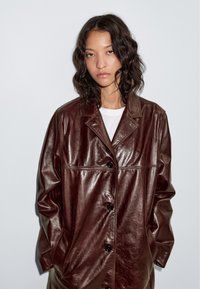 Bruine leren jas met een glanzende textuur, oversized pasvorm, kraagontwerp en zwarte knopen, gedragen over een wit overhemd.