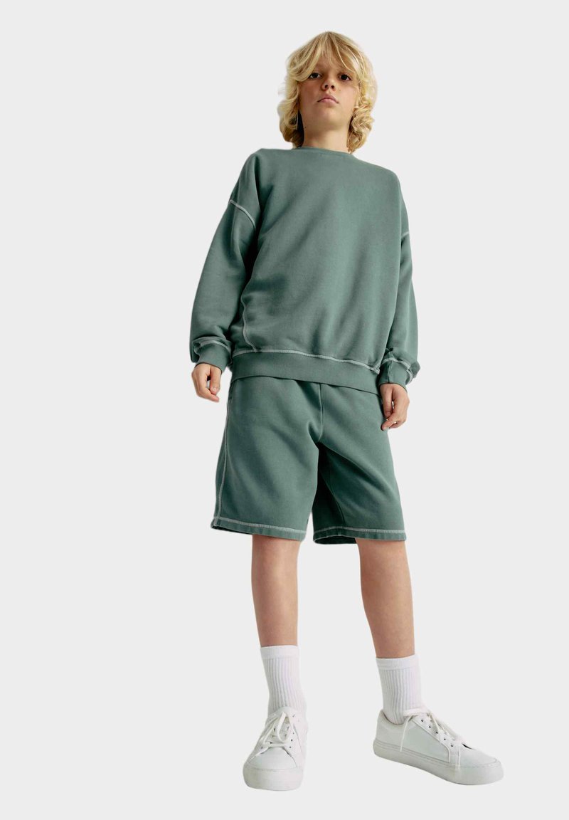 Grøn bomulds sweatshirt og matchende shorts, der har ribbede manchetter og kant. Sammen med hvide sneakers og ribbede hvide sokker.