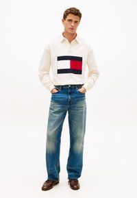 Άσπρο πόλο με μακριά μανίκια και ταμπέλα με λογότυπο σε μπλε ναυτικό και κόκκινο, συνδυασμένο με φαρδιά μπλε τζιν και καφέ δερμάτινα παπούτσια σε απλό φόντο.