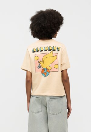 Person with curly hair wearing beige t-shirt featuring a yellow dove holding an olive branch above a globe graphic on the back.