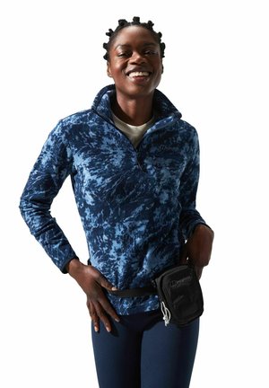 Pull à col zippé en polaire tie-dye bleu avec une poche poitrine zippée. Porté avec un pantalon sombre et un petit sac banane noir.
