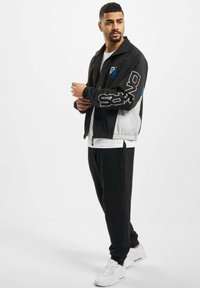 Black and gray windbreaker with a blue accent and white logo on the sleeve, worn over a white t-shirt and paired with black sweatpants.