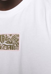 Bílý bavlněný tričko s vyšitým obdélníkovým nášivkem se vzorem olivově zelených a růžových květin a texturovaným povrchem.