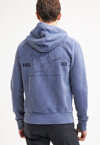 Felpa blu in cotone con tasca frontale e cappuccio regolabile. Presenta un disegno grafico di un personaggio con una chitarra sul retro.