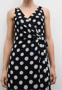 Robe portefeuille noire à pois blancs ; dotée d'un décolleté en V, de fines bretelles et d'un détail noué à la taille. Tissu lisse et léger.