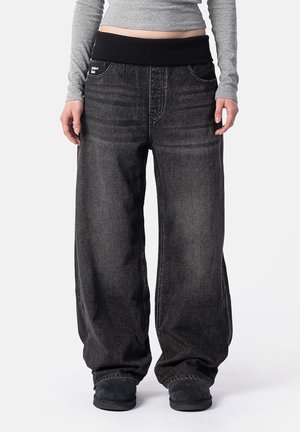 Personne portant un pantalon en jean noir ample lavé avec une large ceinture noire et des chaussures noires sans lacets, se tenant devant un fond uni.