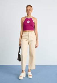 Fialový halter crop top s bielym lemovaním a logom, skombinovaný s béžovými nohavicami so širokými nohavicami a bielymi platformovými sandálmi. Čierna taška na ľavom pleci.