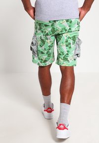 Zielone szorty cargo z motywem liści, z podwójnymi kieszeniami bocznymi i lekkim, przewiewnym materiałem. W połączeniu z białymi trampkami i szarymi skarpetkami.