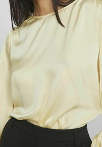 Nainen, jolla on päällään kermaisen värinen satiinipaita, joka on laitettu korkeavyötäröisiin mustiin housuihin, ja suoristettu tumma hius on näkyvissä hartioilla.