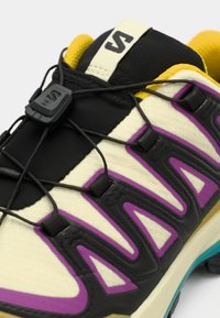 Chaussures de sport avec une tige en maille noire, des accents crème, des éléments de design violets et un col jaune. Elles sont dotées de lacets élastiques et d'une semelle texturée.