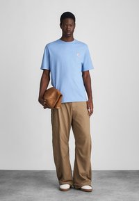 Άνδρας φορώντας ανοιχτό μπλε μπλουζάκι και χαλαρούς καφέ παντελόνες, κρατώντας folded καφέ δερμάτινο μπουφάν, στέκεται μπροστά σε απλό γκρι φόντο.