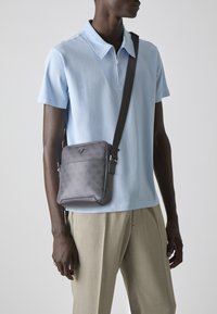 Bolso cruzado gris con un patrón texturizado, que presenta un acento de logo, correa ajustable y bordes suaves, llevado sobre una camiseta polo de color azul claro.