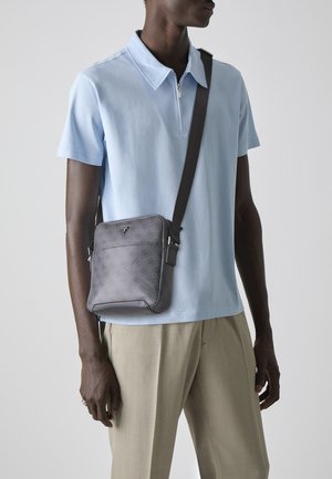 Sac bandoulière gris avec un motif texturé, présentant un accent logo, une sangle ajustable et des bords lisses, porté sur une chemise polo bleu clair.
