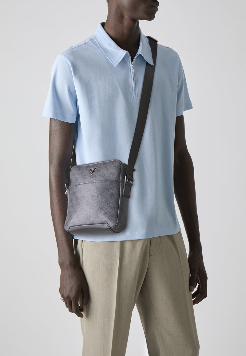 Bolso cruzado gris con un patrón texturizado, que presenta un acento de logo, correa ajustable y bordes suaves, llevado sobre una camiseta polo de color azul claro.