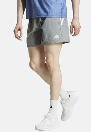 Pantalones cortos deportivos grises con franjas y logo de Adidas en blanco, combinados con zapatillas y calcetines blancos. El material parece transpirable y ligero.