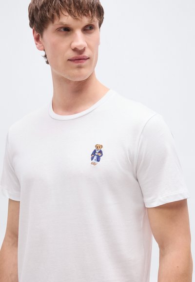 Άσπρο βαμβακερό T-shirt με στρογγυλή λαιμόκοψη και κοντά μανίκια, με ένα μικρό κεντητό αρκουδάκι με μπλε ρούχα στο αριστερό στήθος.