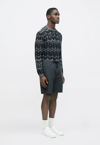Sweter dziergany na drutach w ciemnym odcieniu niebieskiego z wzorem zygzaków, zestawiony z szarymi szortami i białymi tenisówkami. Model stoi w neutralnej pozie na jednolitym tle.