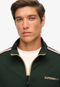 Chaqueta verde con cremallera y detalles en crema, que presenta un cuello alto, tejido acanalado y un logo en el pecho. Cierre delantero con cremallera.
