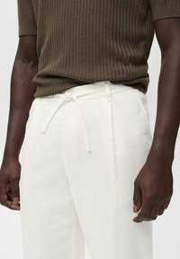 Pantalons en lin blancs avec une taille à cordon de serrage, comportant des poches latérales et une coupe décontractée, associés à un haut en maille côtelée marron.