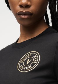 Κοντινό πλάνο ατόμου που φοράει μαύρο πουκάμισο με κυκλικό χρυσό λογότυπο Versace Jeans Couture στο στήθος.