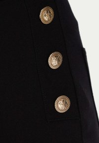 Czarne spodnie z tkaniny z trzema wyraźnymi złotymi guzikami o zaplecionym brzegu. Guziki mają w centrum ozdobny emblemat.