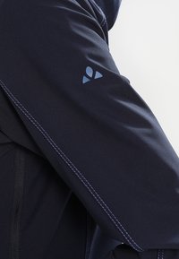 Marinblå jacka med slät yta, kontrasterande ljusa stygn och en liten blå logotyp på vänster ärm.