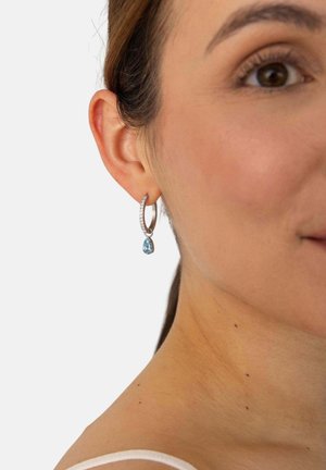 Nahaufnahme eines Frauenohres mit einem silbernen Creolen-Ohrring mit kleinen Steinen und einem hängenden blauen tropfenförmigen Edelstein.