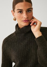 Dunkles, gestricktes Rollkragensweatshirt mit gerippter Textur. Das Model hat eine Hand im Gesicht und trägt goldene Creolen, die eine neutrale Farbpalette präsentieren.