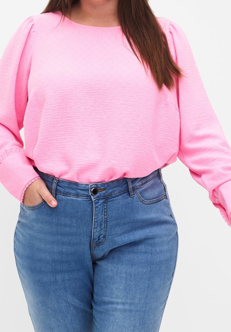 Blouse longue à manches longues texturée rose avec épaules bouffantes, associée à un jean denim bleu taille haute, doté d'une poche avant et d'une fermeture à boutons.