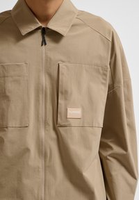 Lichtbruine zip-up jas van stof met een gladde textuur, voorzien van twee voorzakken en een merkpatch op de linkerborst.