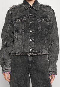 Svart denimjacka med en kortad design, fransat fåll och knäpps med knappar. Har två bröstfickor och texturerade axelpartier.