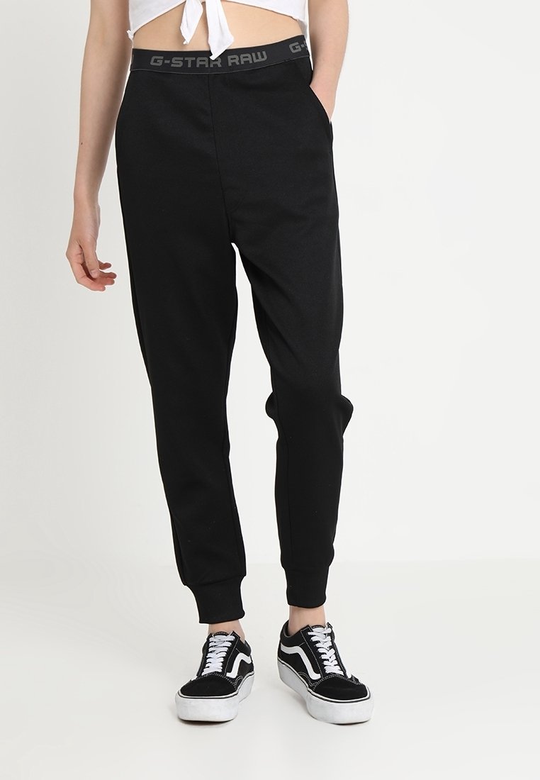 Personne portant un pantalon de jogging noir G-Star Raw avec une taille élastique et des baskets noires et blanches, debout devant un fond blanc uni.