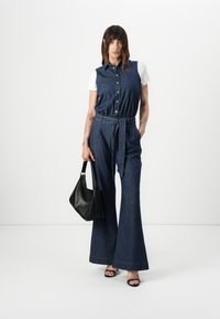 Denim jumpsuit met een knoopsluiting aan de voorkant, een strik taille en brede uitlopende benen. Met een kraag en een lichte katoenen onderlaag. Zwarte handtas.